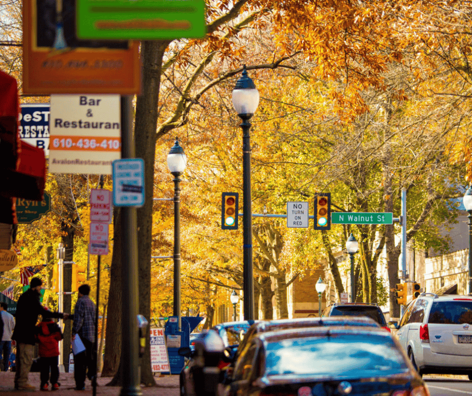 A vibrant street scene in Philadelphia during fall, with colorful autumn leaves on trees, pedestrians walking on the sidewalk, and cars stopped at a traffic light. Street signs, storefronts, and a warm urban atmosphere are visible.