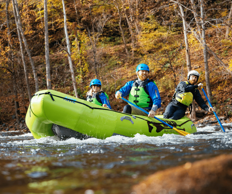 Three people whitewater rafting on a river in Erwin, Tennessee with Blue Ridge Paddling. They are wearing helmets and life jackets in a bright green inflatable raft, surrounded by autumn trees with colorful foliage.