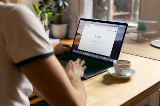 Person using a laptop with the Google search page open, sitting at a wooden table with a cup of coffee. Promotes an educational video for small businesses about important search updates that impact how customers find local businesses online.