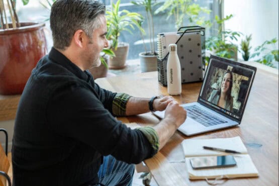 A man sits at a wooden desk having a video consultation on a laptop; an open notebook, pen, phone, and water bottle are nearby.